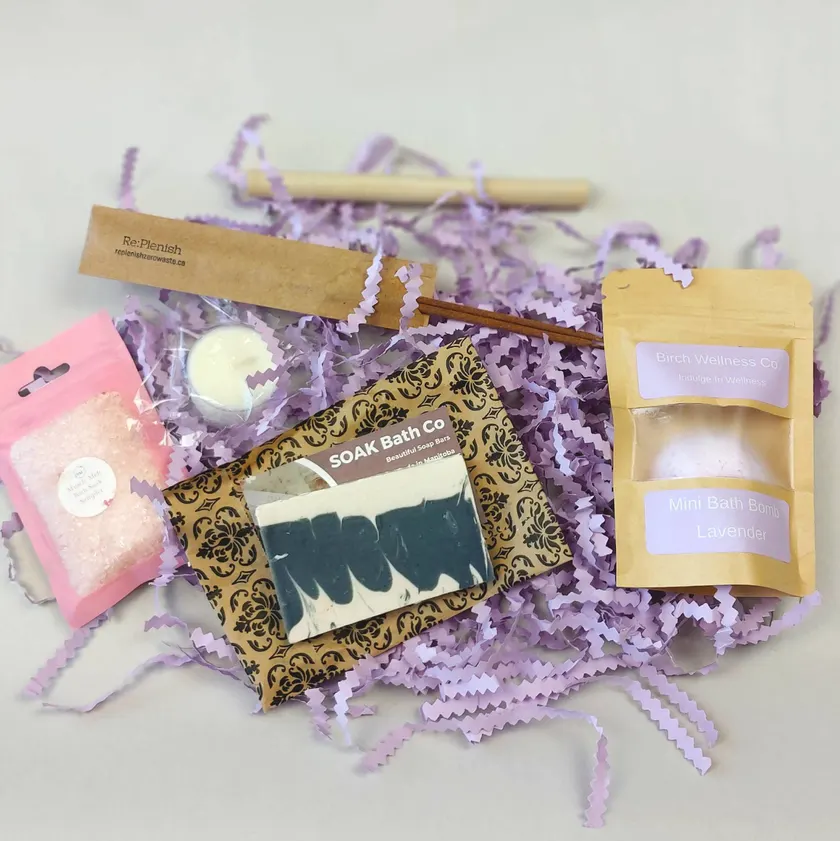 Bath extras arranged on lavender crinkle paper: a bath salt sampler, a tealight candle, incense sticks with a wooden holder, a marbled soap bar, and a lavender mini bath bomb.