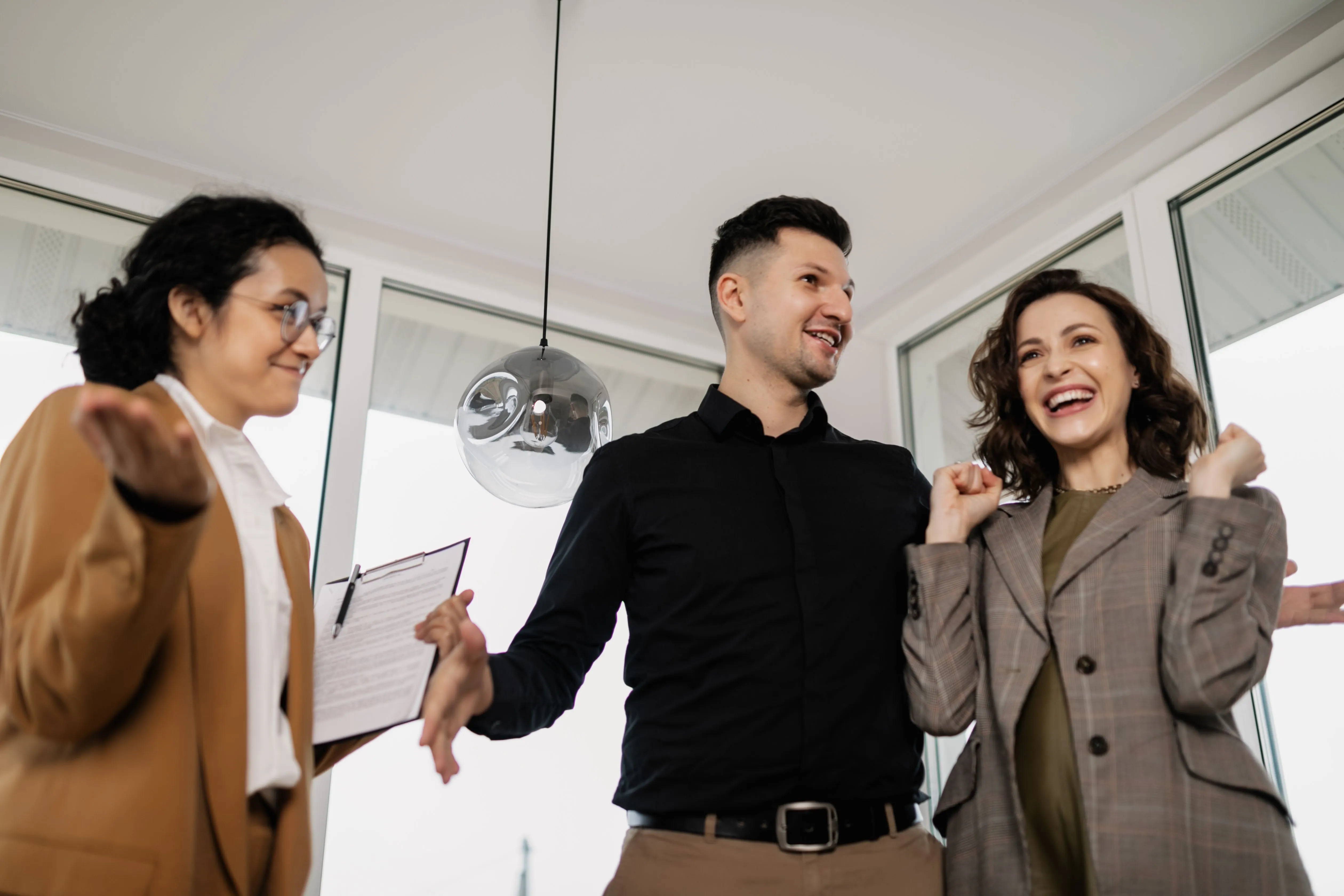A real estate agent gesturing across a bright, empty living room while a smiling couple takes it in.
